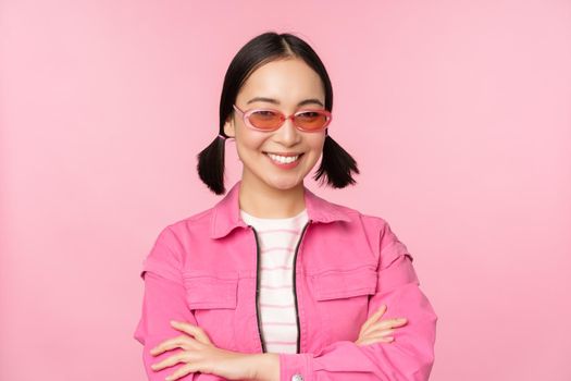 Stylish asian girl in pink clothes and sunglasses, smiling and looking happy at camera, standing over studio background.