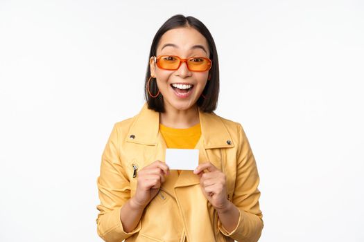 Portrait of beautiful modern asian girl in sunglasses, smiling happy, showing credit card, standing over white background. copy space