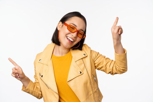 Smiling asian brunette woman in sunglasses, pointing fingers sideways, left and right, showing variants, laughing and dancing, wearing sunglasses, white background.