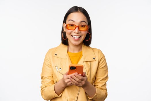 Modern asian girl in sunglasses using her mobile phone, smiling and looking happy, posing against white background. Copy space