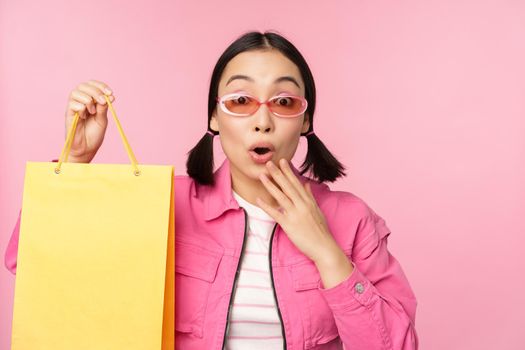 Shopping. Stylish asian girl in sunglasses, showing bag from shop and smiling, recommending sale promo in store, standing over pink background.