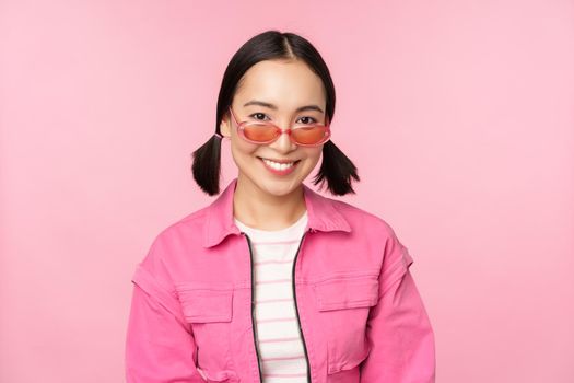 Close up of beautiful asian female model in stylish sunglasses, posing against pink background in trendy outfit, copy space.