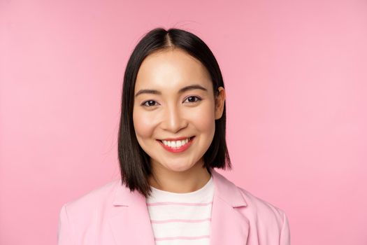 Close up portrait of asian corporate woman, looking professional, smiling at camera, wearing suit, standing over pink background. Copy space