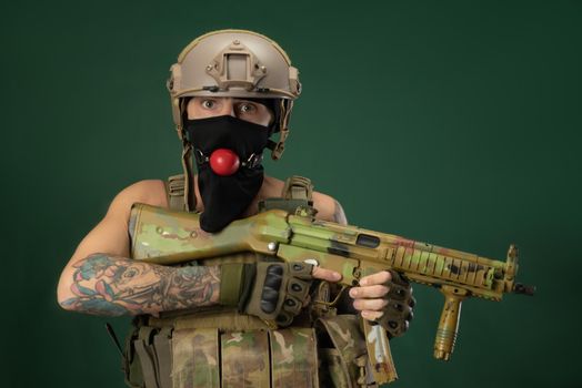 male soldier in military clothes, a helmet with a bdsm gag in his mouth expresses emotions holding an automatic rifle in his hands, photo joke