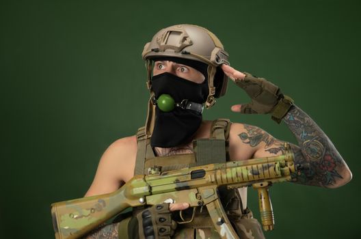 male soldier in military clothes, a helmet with a bdsm gag in his mouth expresses emotions holding an automatic rifle in his hands, photo joke