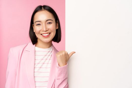 Portrait of smiling asian businesswoman in suit, corporate lady pointing finger at white empty wall, board with info or advertisement, standing over pink background.