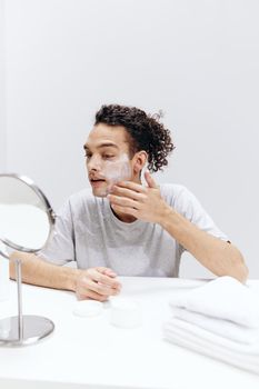 guy with curly hair puts cream on the face but sitting at the table in front of the mirror isolated background. High quality photo