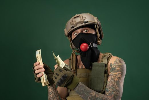 soldier man in military clothes helmet with a bdsm gag in his mouth expresses emotions holding money dollars in his hands, photo joke