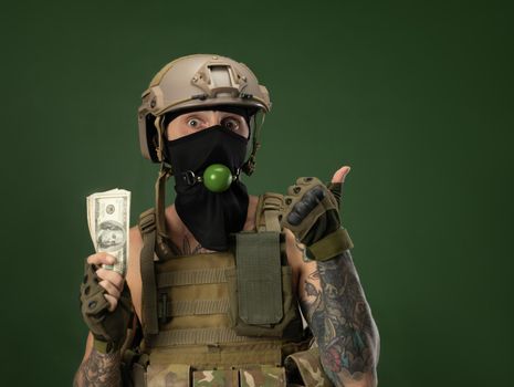 soldier man in military clothes helmet with a bdsm gag in his mouth expresses emotions holding money dollars in his hands, photo joke