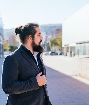 Young businessman with long hair and beard standing minding his own business during his work break, on the street next to the offices with. vertical photo on a sunny and clear day. Size view