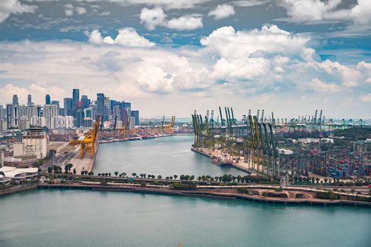 The huge busiest logistic port in Singapore, plenty of cranes to move containers, huge cargo ships in the background, skyscrapers on background. High quality photo