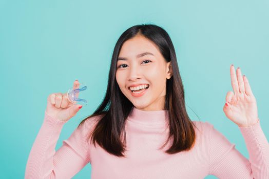 Portrait Asian beautiful young woman smiling holding silicone orthodontic retainers for teeth and show finger brackets saying OK sign, isolated on blue background, Dental hygiene healthy care concept