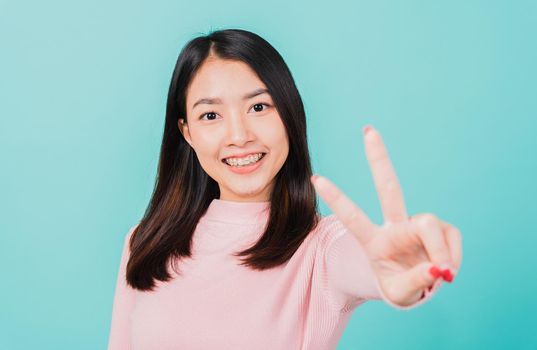 Young Asian beautiful woman smiling wear silicone orthodontic retainers on teeth showing v-sign victory finger isolated on blue background, retaining after removable braces. Dental hygiene concept