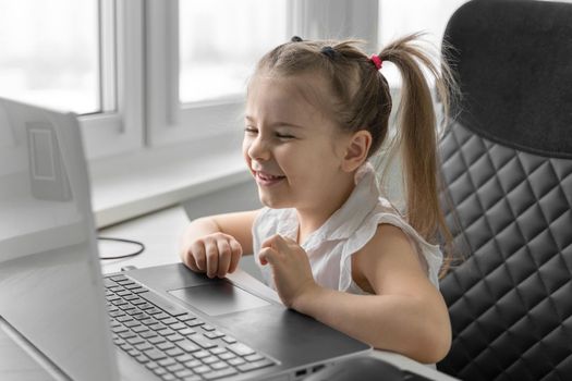 cute little caucasian girl sits at table, watching at laptop screen