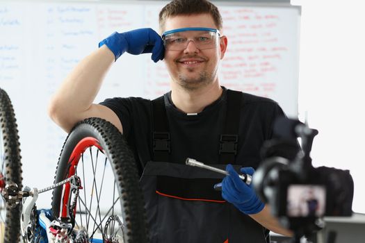 Portrait of happy handyman try to fix broken bicycle, film process on videocamera, mechanic start online channel. Blogger, content, hobby, creative concept