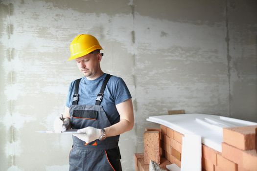 Portrait of concentrated male worker hold clipboard and write down needed work material for renovation. Foreman in uniform and helmet. Construction concept