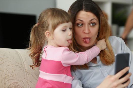 Portrait of mother and daughter show tongues on camera, having fun taking selfie on smartphone. Girl and woman enjoy time together. Family time concept