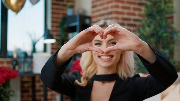Sexy blonde woman showing heart shape sign on camera to celebrate valentines day holiday with flowers and presents. Attractive beautiful person advertising romance and love symbol.