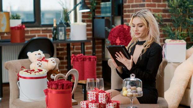 Elegant blonde woman smiling at tablet computer while talking with remote boyfriend during valentine day surprise. Attractive cute girlfriend enjoying romantic gift advertising romance anniversary