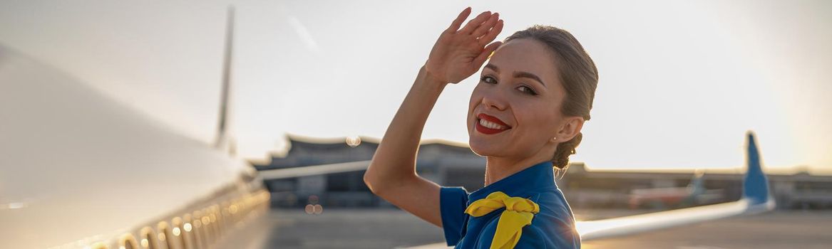 Portrait of cheerful air stewardess in blue uniform smiling at camera, posing outdoors at the sunset. Commercial airplane near terminal in an airport in the background. Aircrew, occupation concept