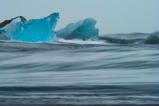 Spectacular blue icebergs over the black sand beach beaten by the breaking waves of the ocean, long exposure