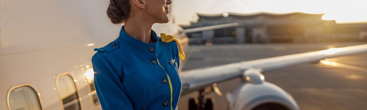 Thoughtful air stewardess in blue uniform looking away, standing outdoors at the sunset. Commercial airplane near terminal in an airport in the background. Aircrew, occupation concept