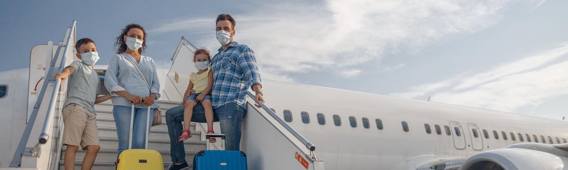 Happy family in protective masks, parents with two little children standing on airstair, boarding the plane in the daytime. People, traveling, vacation concept