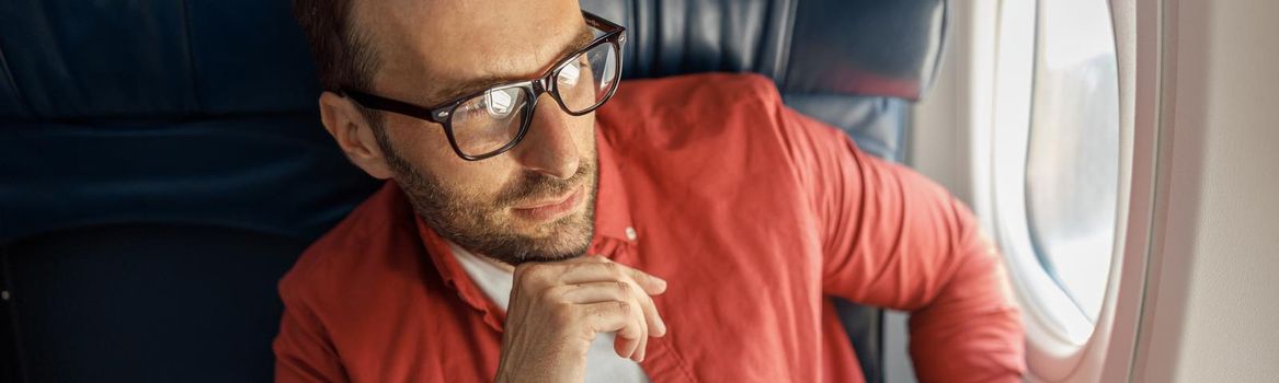 Handsome caucasian man in casual wear and glasses looking thoughtful, sitting on the plane near the window. Relax, travel, vacation, transportation concept