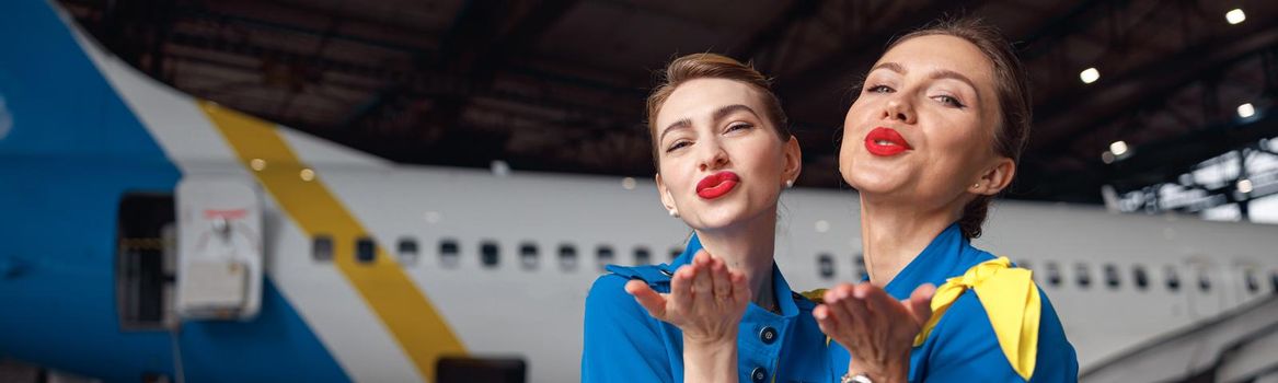 Two air stewardesses in stylish blue uniform blowing kisses to camera, standing together in front of passenger aircraft in hangar at the airport. Occupation concept