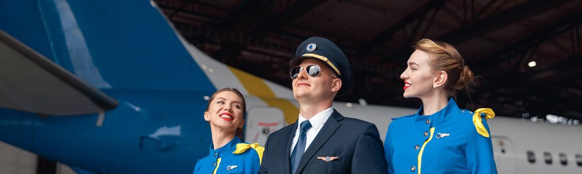 Happy pilot in uniform and aviator sunglasses walking together with two air stewardesses in blue uniform in front of big passenger airplane in airport hangar. Aircraft, occupation concept