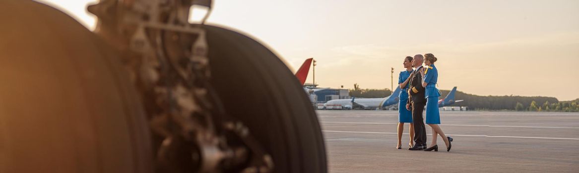 Full length shot of male pilot posing for photoshoot together with two air hostesses in blue uniform, standing in an airport terminal at sunset. Aircraft, aircrew concept