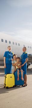 Full length shot of beautiful stewardesses standing near two kids sitting on their luggage in front of big airplane and smiling at camera. Family, traveling, vacation concept