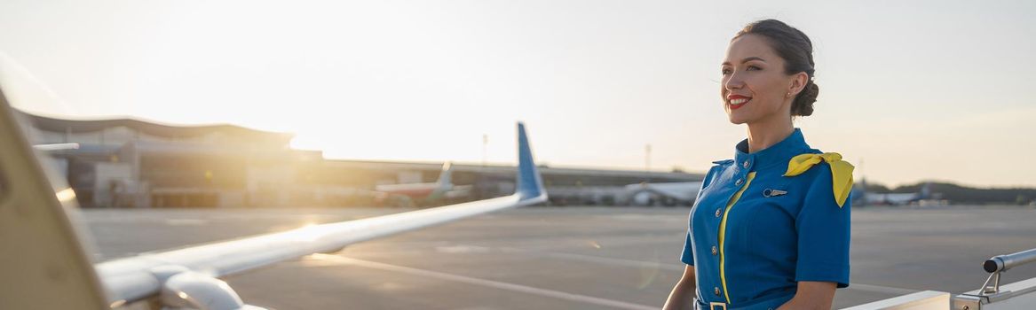 Pretty air stewardess in blue uniform smiling away, standing outdoors at the sunset. Commercial airplane near the terminal in an airport in the background. Aircrew, occupation concept