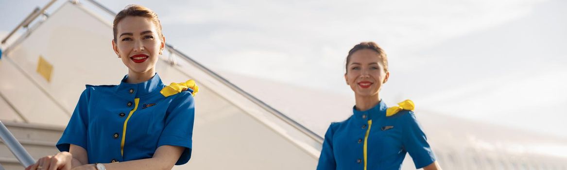 Two beautiful air stewardesses in blue uniform smiling at camera, standing on airstair and welcoming passengers. Aircrew, occupation concept