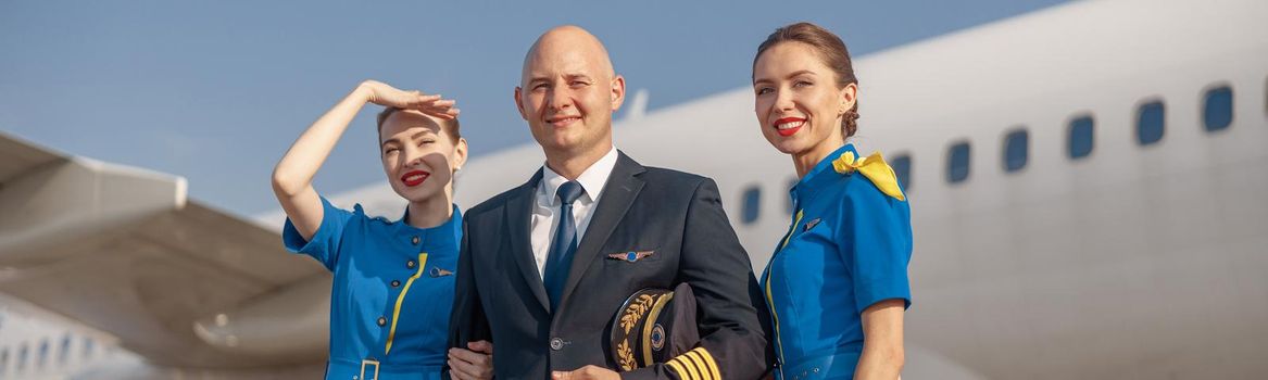 Happy pilot and two attractive stewardesses standing together in front of an airplane and smiling after landing. Aircraft, aircrew, occupation concept