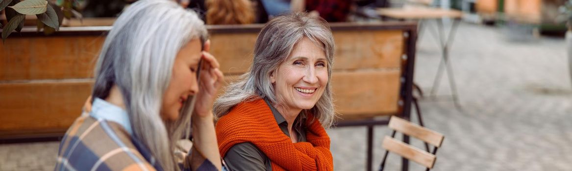 Happy mature woman with grey haired friend rest together sitting at small wooden table in street cafe on nice autumn day