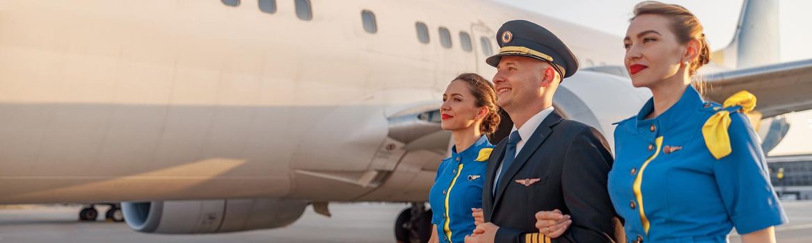 Excited male pilot walking together with two flight attendants in blue uniform in front of an airplane in terminal at sunset. Aircraft, aircrew concept