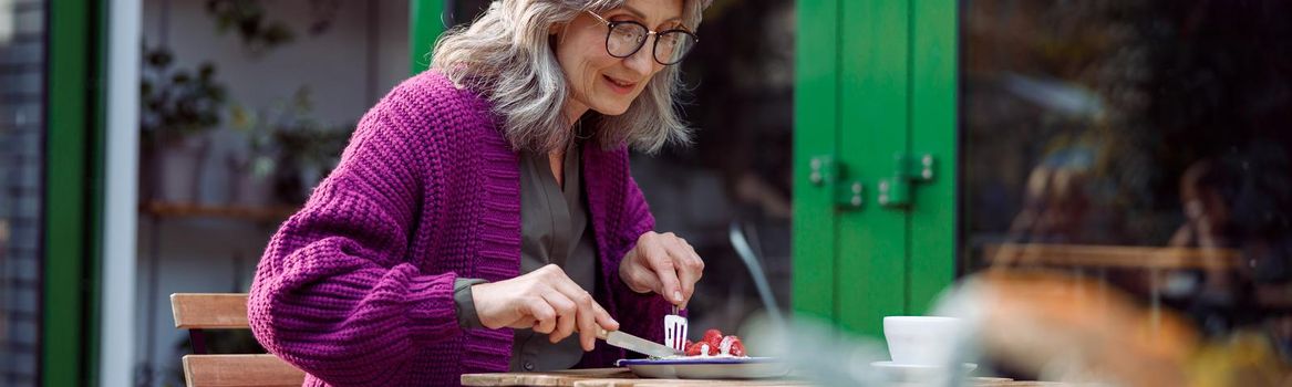 Happy mature woman with glasses in purple jacket eats delicious dessert at table on outdoors cafe terrace on autumn day