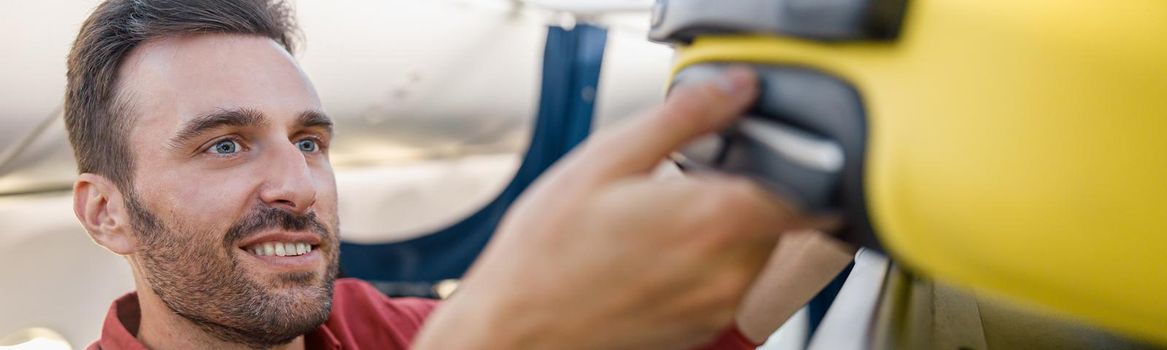 Portrait of cheerful man pulling out suitcase from hand luggage compartment, overhead locker while traveling by plane. Vacation, transportation concept