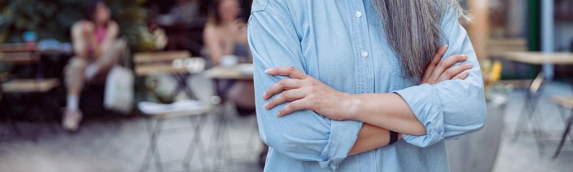 Positive hoary haired senior Asian lady with crossed arms in blue blouse stands against outdoors cafe terrace on nice autumn day