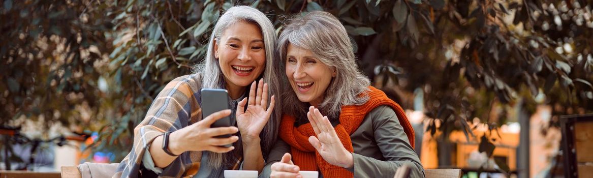 Positive mature women friends wave hands at videochat via smartphone sitting at small table in street cafe on nice autumn day