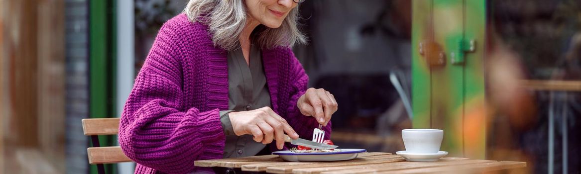Pretty grey haired senior lady in purple knitted jacket eats delicious dessert sitting at table on outdoors cafe terrace on autumn day