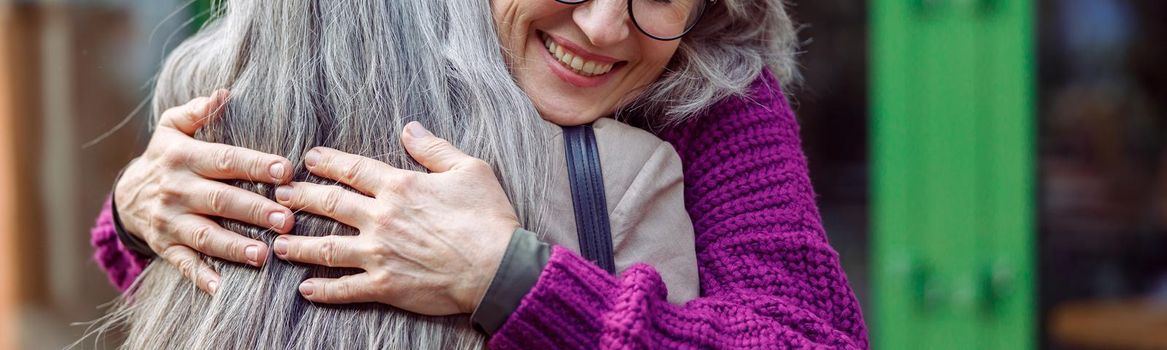 Happy senior woman with glasses in knitted jacket hugs best friend with long silver hair meeting on city street