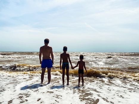 A young father with two sons are standing on the seashore among the waves and looking at the horizon enjoying the sea breeze