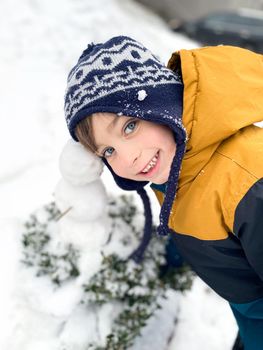 Portrait of a happy five year old boy in the snow in winter. The boy plays and poses with a little snowman