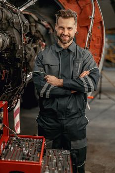 Joyful bearded man maintenance technician holding wrench and smiling while standing near airplane at repair station