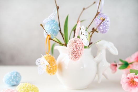 Home interior with easter decor. Vase with willow tree branches with Easter eggs and bunny on white table and background with copy space.