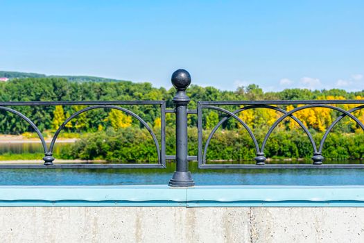 Black decorative fence on the embankment against the background of the river and the forest. Transparent metal fence