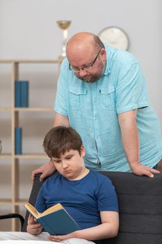 A son sits on the sofa and reads his school reading and the father supervises by standing behind him. The father is helping with his son's studies.