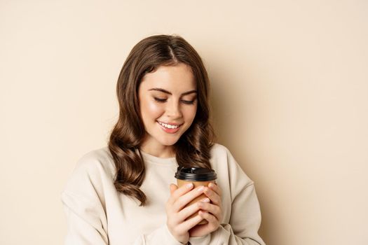 Beautiful authentic woman smiling, holding warm cup of coffee and looking happy, standing over beige background.